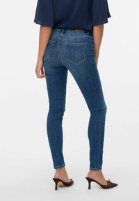 Jeans de mezclilla azul ajustados con bolsillos traseros, lavado medio, textura suave, llevados con mule de tacón marrón, vista desde la parte trasera.