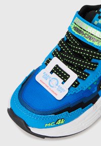 Blauwe sportieve schoen met een bovenwerk van zwart en groen synthetisch gaas, voorzien van een adaptieve sluiting en een textuurzool. Accenten met "MC4K."