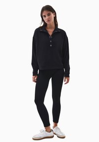 Suéter negro con un cuello alto y cierre de botones, combinado con mallas negras y zapatillas blancas con suelas marrones.