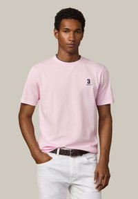 Giovane uomo che indossa una t-shirt rosa chiaro a maniche corte con un piccolo logo "3 Hackett" sul petto, pantaloni bianchi e una cintura intrecciata marrone.