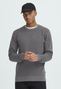 Szürke kötött pulóver kerek nyakkal, bordás mandzsettákkal és hem. Fehér ing felett viselve. A modell fekete sapkát visel. Sima textúra, minta nélkül.