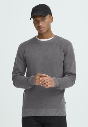 Grijze gebreide trui met een ronde hals, geribde manchetten en zoom. Draagt over een wit shirt. Model draagt een zwarte pet. Gladde textuur, geen patronen.