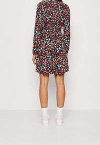 Schwarzes Blumenkleid mit roten und blauen Blumen, langen Ärmeln, tailliertem Design und schwingendem Rock. Kombiniert mit weißen Sneakers und Socken.