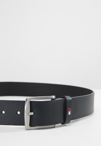 Cinturón de cuero negro con una hebilla rectangular plateada. Presenta pequeños agujeros para ajustes y un acento lateral en rojo y blanco.