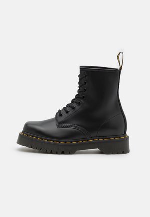 Bottines en cuir noir avec un bout arrondi, un design robuste à lacets, des coutures jaunes, une semelle en caoutchouc texturée et une languette à l'arrière.