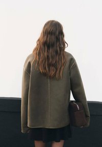 Felpa oversize verde oliva con spalle scese e taglio dritto, abbinata a una gonna scura e con una borsa marrone.