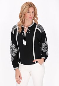 Blusa nera con ricami floreali bianchi, dettagli in pizzo e scollatura a fiocco. Maniche lunghe con motivi dettagliati, abbinata a pantaloni bianchi.