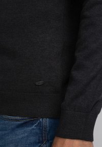 Dark gray sweater with a ribbed hem and cuffs, featuring a small black logo tag near the bottom. Material appears soft and textured.