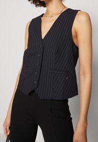 Kvinna klädd i en svart randig ärmlös skjortväst som matchar med svarta tajta byxor, ett tunt guldhalsband och en guldring på vänster hand.