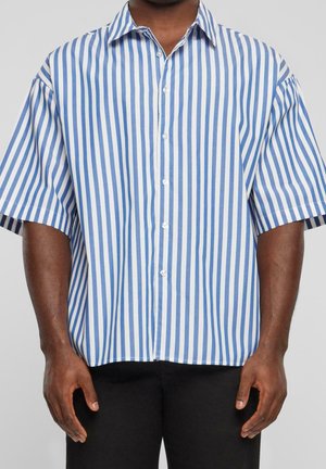 Mann trägt kurzärmliges, blau-weiß gestreiftes Hemd mit schwarzen Hosen vor grauem Hintergrund.