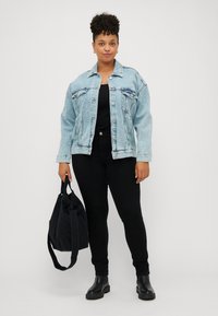 Kvinne med krøllete hår iført en lys blå denimjakke, svart topp, svarte bukser og svarte støvler, som holder en svart totebag med den ene hånden.