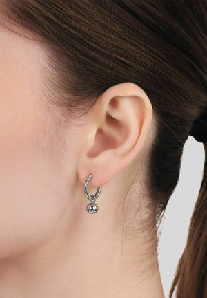 Nahaufnahme eines Frauenohres mit dunkelbraunem, zurückgebundenem Haar, das einen kleinen silbernen Creolen-Ohrring mit einer hängenden silbernen Kugel trägt.