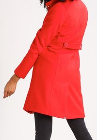 Manteau rouge au design structuré, comprenant une ceinture et des détails de boutons, manches longues et texture lisse. Porté avec un pantalon noir.