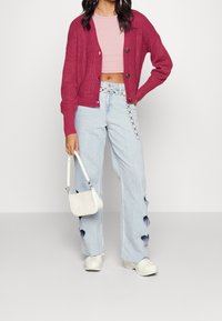 Cotton knit cardigan in burgundy with buttons, pink crop top, light blue jeans featuring heart-shaped cutouts, white bag, and chunky sneakers.