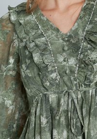 Vestido floral verde con escote volado, detalles de botones y una cinta en la cintura. La tela texturizada presenta patrones sutiles y un acabado brillante.