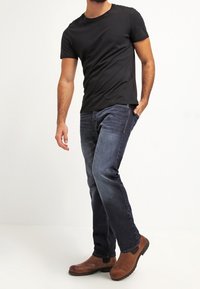 Camiseta negra de manga corta, vaqueros azul oscuro y botas de tobillo marrones. El ajuste es casual, con un diseño de pierna recta y un detalle mínimo.
