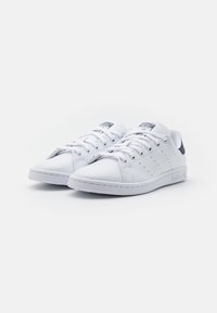 Vita lädersneakers med platta snören, perforeringar på sidorna och en kontrasterande häl. Har en logotyp på tungan och vadderad innersula.