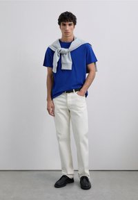 Giovane uomo che indossa una maglietta blu, pantaloni bianchi, scarpe nere e una felpa grigia legata attorno alle spalle, in piedi su un pavimento grigio.