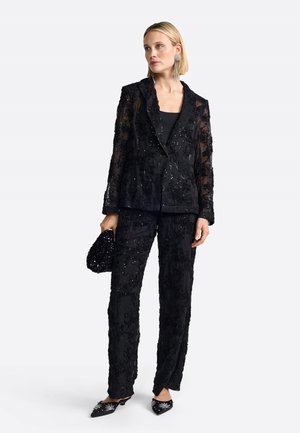 Schwarzer transparenter Blazer und passende Hose, verziert mit floralen Stickereien und Pailletten. Zubehör umfasst eine bestickte Clutch und verzierte Schuhe.