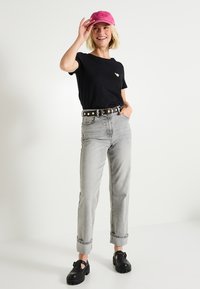 Camiseta negra de manga corta con emblema de corazón, jeans gris de tiro alto, gorra rosa y zapatos negros gruesos, de pie frente a un fondo simple.