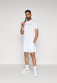 Witte sport t-shirt met een klein logo, gecombineerd met witte shorts en sportschoenen. Houdt een tennisracket vast. Eenvoudig ontwerp, geen patronen.
