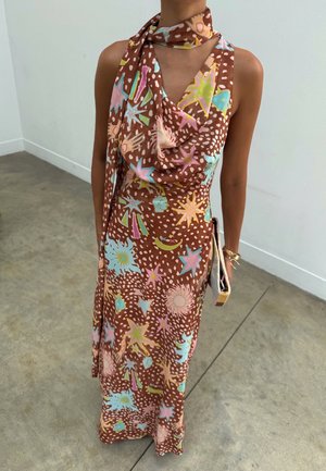 Femme portant une robe marron sans manches et une écharpe ornée de motifs colorés d'étoiles, de soleil et de lune, tenant une pochette beige et un bracelet doré.