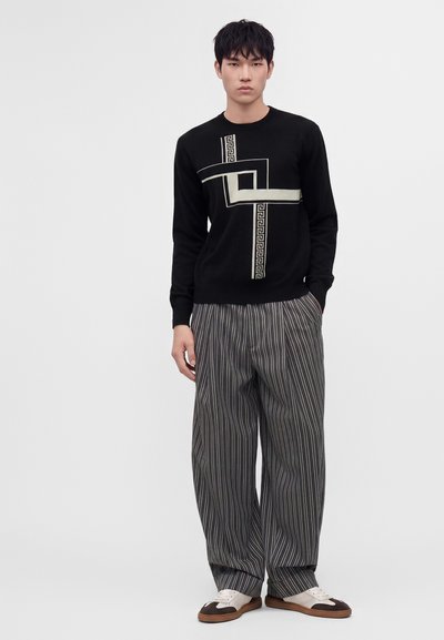 Homme portant un pull noir avec un motif géométrique blanc, un pantalon large gris à fines rayures et des chaussures bicolores marron et blanc, debout les mains dans les poches.