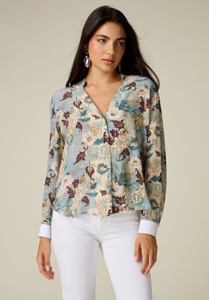 Frau mit langen dunklen Haaren, die große geometrische Ohrringe, eine florale Bluse mit Knöpfen in Blau-, Creme- und Brauntönen sowie weiße Hosen trägt.