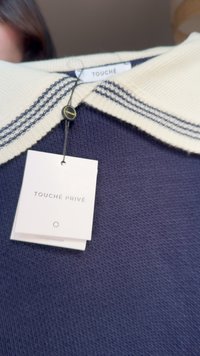 Pulover tricotat bleumarin cu un guler crem cu dungi bleumarin. Eticheta spune "TOUCHÉ PRIVÉ." Țesătura are un aspect texturat și respirabil.