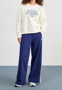 Krämfärgad sweatshirt med texten "Adidas Originals" i blått, parat med löst sittande marinblå byxor med gula sidostråk. Texturerad tyg synlig.