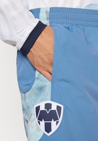 Los pantalones cortos de color azul claro cuentan con un bolsillo lateral y un logo en forma de escudo blanco con detalles en azul marino. La tela tiene una textura suave y ligeramente brillante.