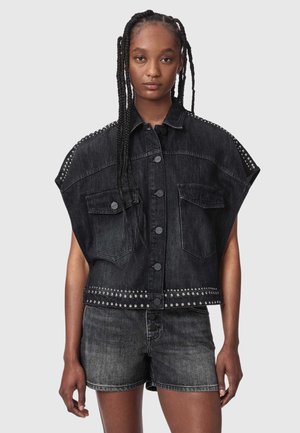 Jonge persoon met lange vlechten, gekleed in een mouwloze zwarte spijkerjas met studs en bijpassende zwarte spijkershorts tegen een effen achtergrond.