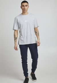 Grau meliertes T-Shirt mit kurzen Ärmeln und kleinem weißen Logo auf der Brust, kombiniert mit marineblauen Jogginghosen und schwarzen Sneakers.
