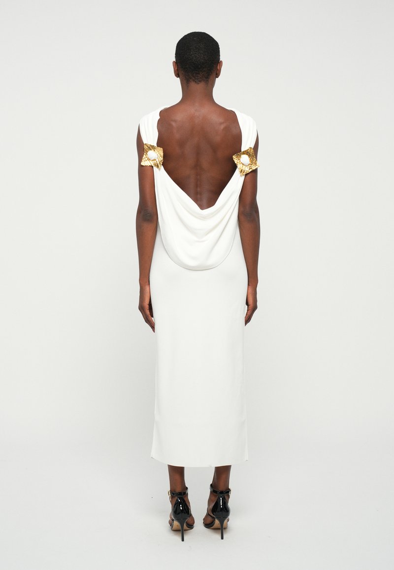 Robe blanche drapée avec un dos profond, ornée de délicates accentuations en métal doré au niveau des épaules. Texture lisse et silhouette ajustée.