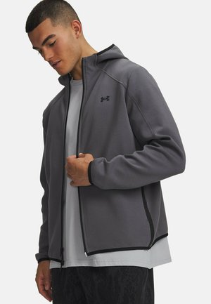 Man draagt een grijze zip-up hoodie met capuchon en zwarte rand over een lichtgrijs shirt, kijkt naar beneden terwijl hij de jas gedeeltelijk dichtritst.