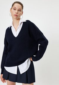 Merinotuulepaita tummansinisellä värillä, V-pääkohdalla, ribbytekstuuri ja pitkät hihat, kerros valkoisen kauluspaitaan ja pinstrioituneeseen liukuvaan hameeseen.