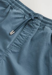 Pantalones de baño azules con cintura elástica, cierre de cordón y un bolsillo en el lado izquierdo. Tejido suave con un ligero brillo.