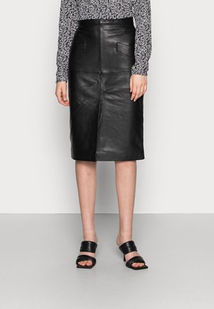 Femme portant une jupe en cuir noire longueur genou avec poches avant, un haut à manches longues à motif et des sandales noires à talons ouvertes.
