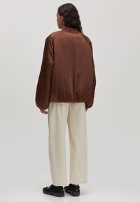 Bruine satijnen bomberjack met raglanmouwen, elastische tailleband en minimalistisch ontwerp. Gecombineerd met crèmekleurige wijde broek en zwarte sneakers.