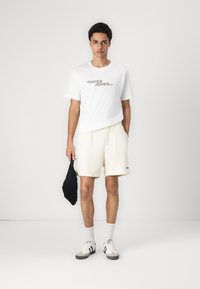 Camiseta blanca con logo gris, pantalones cortos crema y zapatillas grises con rayas negras. Sosteniendo un pequeño bolso negro, de pie contra un fondo sencillo.