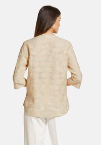 Beige strukturierte Bluse mit einem dezenten Muster, Dreiviertelärmeln und Rundhalsausschnitt, kombiniert mit weißen Hosen.