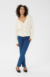 White cardigan made from soft material, featuring a V-neck, ribbed cuffs, and wooden buttons, paired with blue fitted jeans and black shoes.