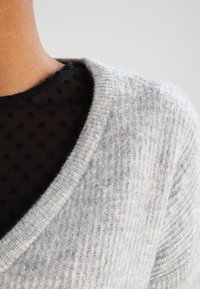 Maglione grigio a maglia con un motivo testurizzato e scollatura a V, indossato sopra un top in rete nera con pois. Tessuto morbido e peloso con punti di cucitura visibili.