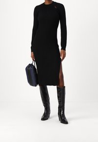 Abito lungo nero a coste con maniche lunghe, scollatura alta e spacco laterale, abbinato a stivali neri lucidi fino al ginocchio e a una borsa blu scuro.