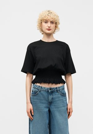 Kvinde med kort krøllet blondt hår iført en sort cropped T-shirt med flæsekant og højtaljede, løstsiddende blå jeans mod en ensfarvet baggrund.