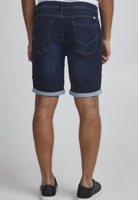 Shorts en denim bleu avec un ourlet retroussé, comprenant deux poches arrière et des coutures jaunes contrastantes. Portés avec des baskets noires.