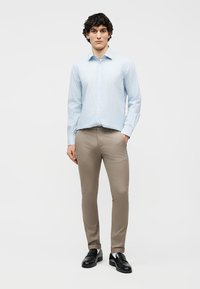 Νεαρός άνδρας που στέκεται φορώντας ελαφρύ μπλε ριγέ πουκάμισο, μπεζ παντελόνι στενής γραμμής και μαύρα δερμάτινα loafers σε απλό φόντο.