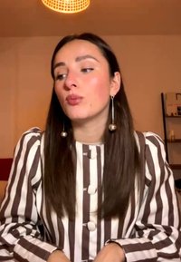 Jeune femme aux longs cheveux bruns portant un chemisier rayé marron et blanc et des boucles d'oreilles sphériques pendantes, regardant vers sa gauche à l'intérieur.