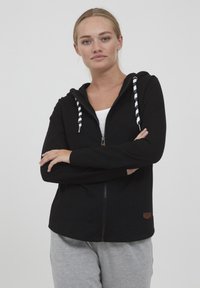 Juodas zip-up megztinis su tekstūruotu paviršiumi, baltais ir juodais dryžuotais raišteliais ir mažu ruduo logotipu apatinėje dalyje.