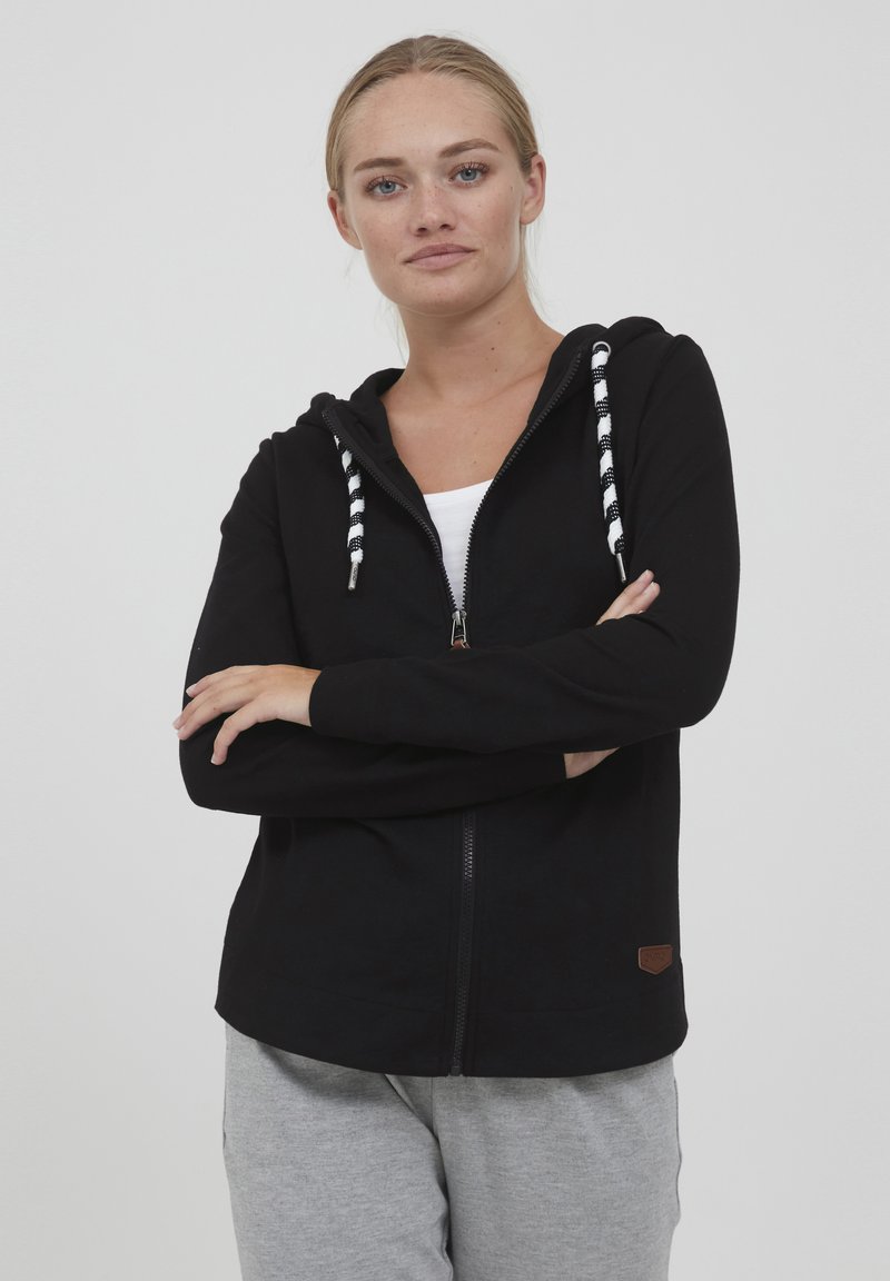 Juodas zip-up megztinis su tekstūruotu paviršiumi, baltais ir juodais dryžuotais raišteliais ir mažu ruduo logotipu apatinėje dalyje.